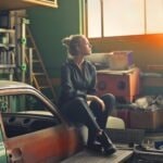 Junge Frau in einer Werkstatt, auf einer Motorhaube sitzend.